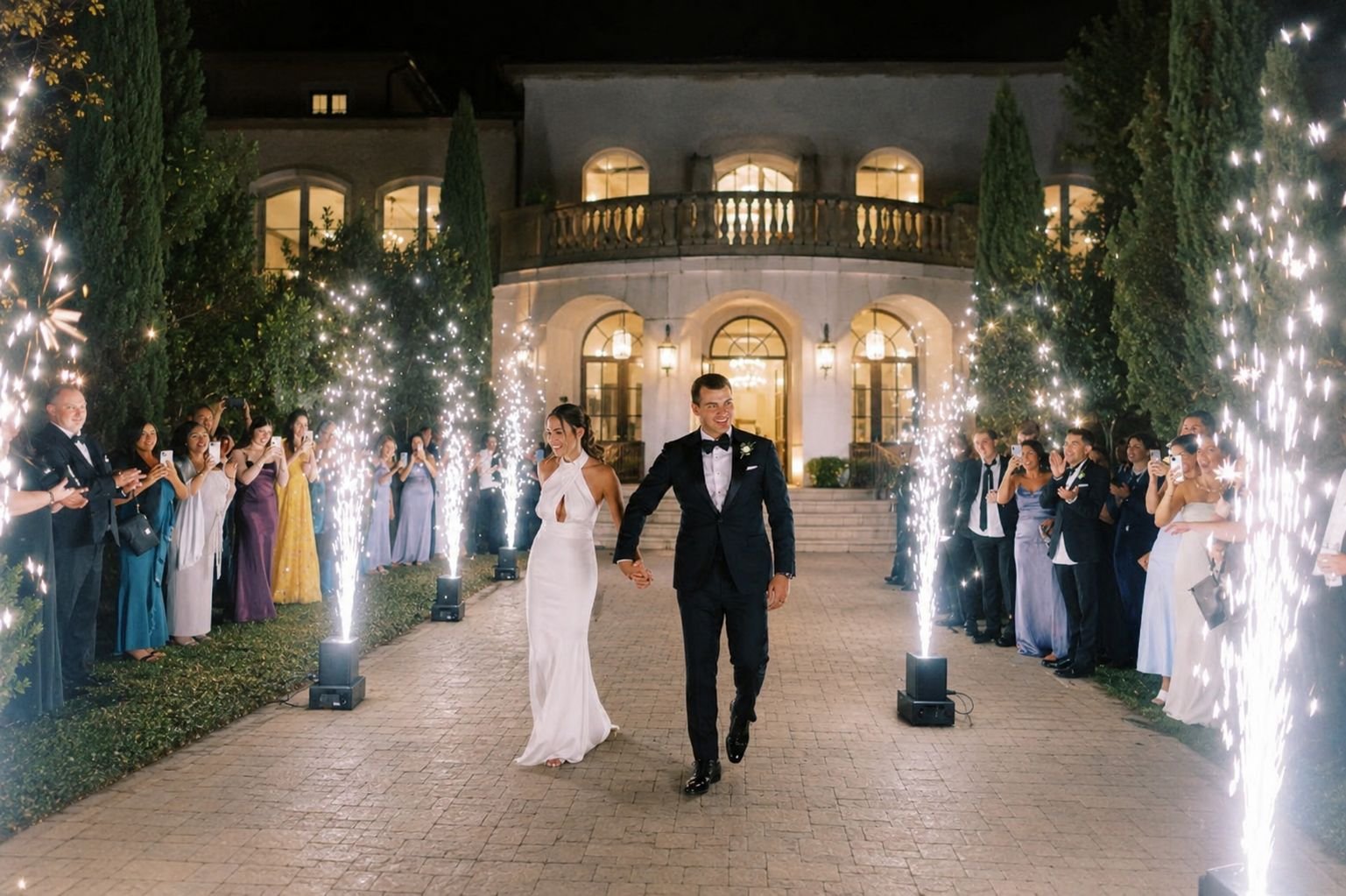 A newlywed couple in wedding attire walking hand in hand through fireworks and sparklers on a brick pathway outside a large, illuminated house at night, with wedding guests taking photos.