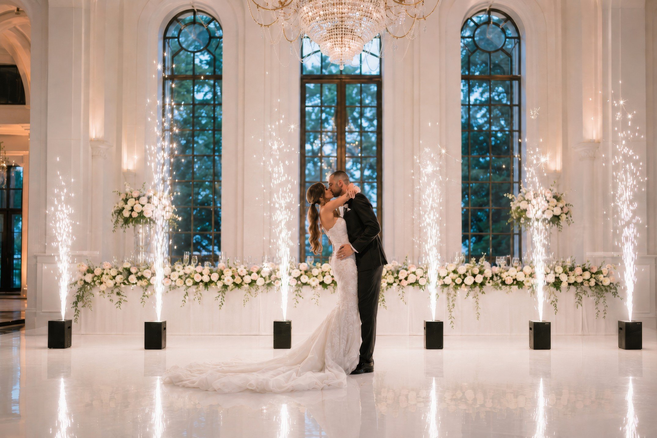A bride and groom share a kiss on their wedding dance floor in an elegant indoor venue with tall arched windows, a grand chandelier, and floral decorations, during their wedding celebration.