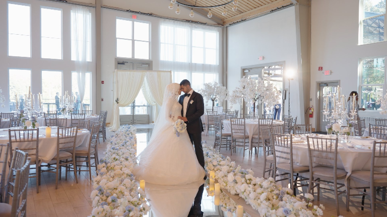 A bride and groom standing close in a decorated wedding reception hall, with floral arrangements and candles on tables.