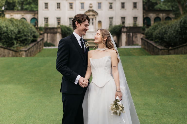 A bride and groom standing on a green lawn, gazing at each other during their wedding celebration in front of a historic white building.
