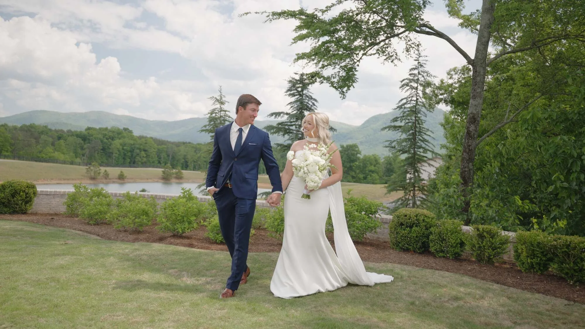 A happy bride and groom holding hands and walking outdoors on a lush green lawn with trees, hills, and a lake in the background on a cloudy day.