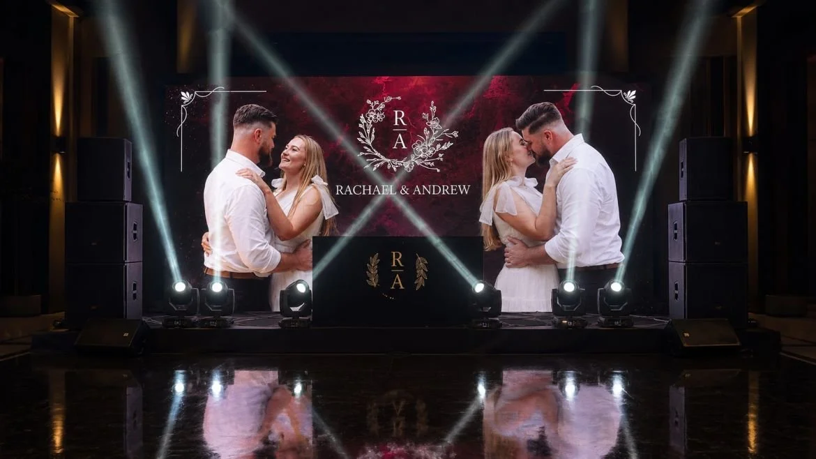 Two couples dancing and kissing at a wedding reception with a large LED display showing their names, Rachael & Andrew, and a monogram with the initials R and A.