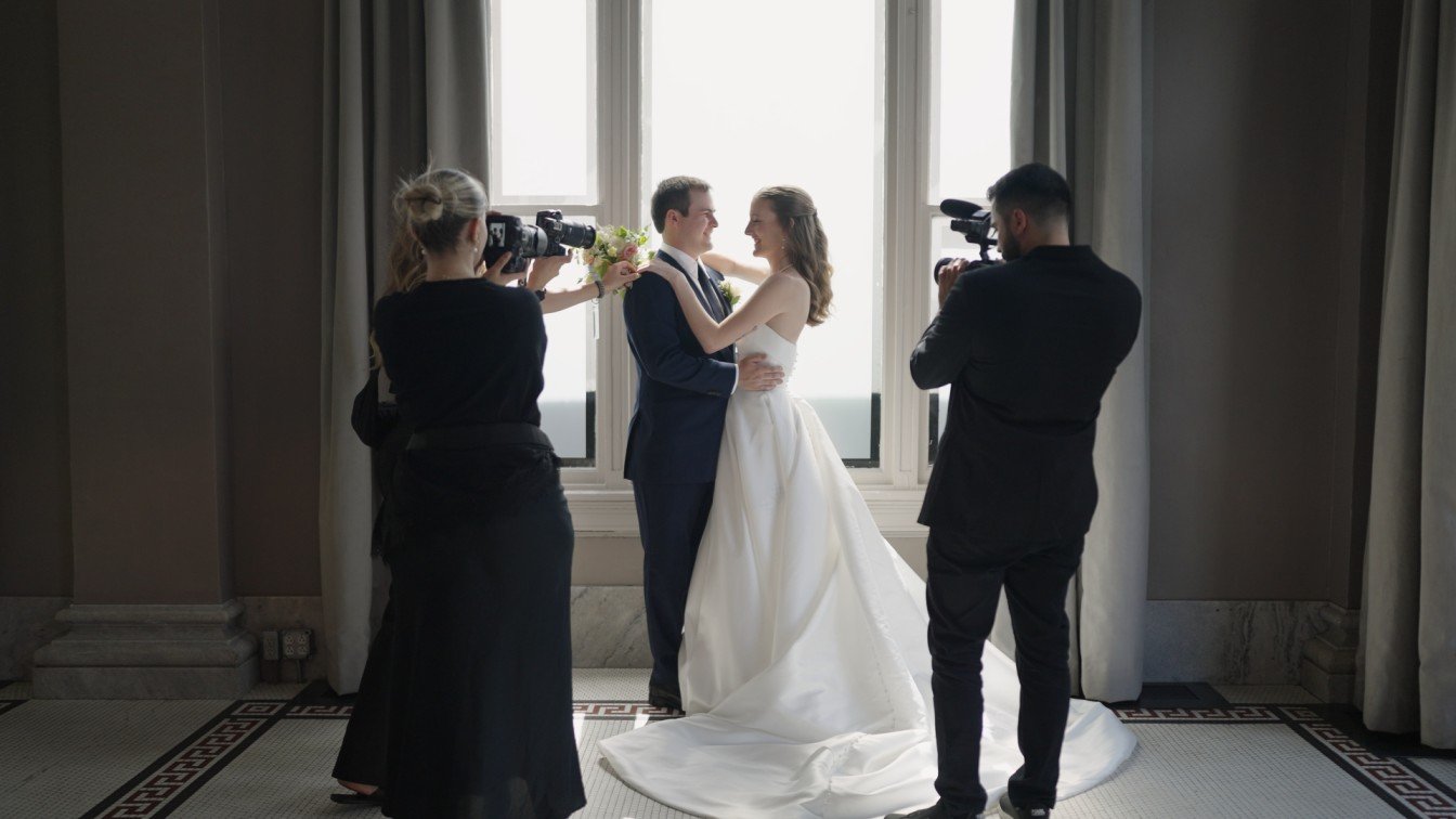 A bride and groom holding each other while being filmed and photographed during their wedding inside a room with large windows.