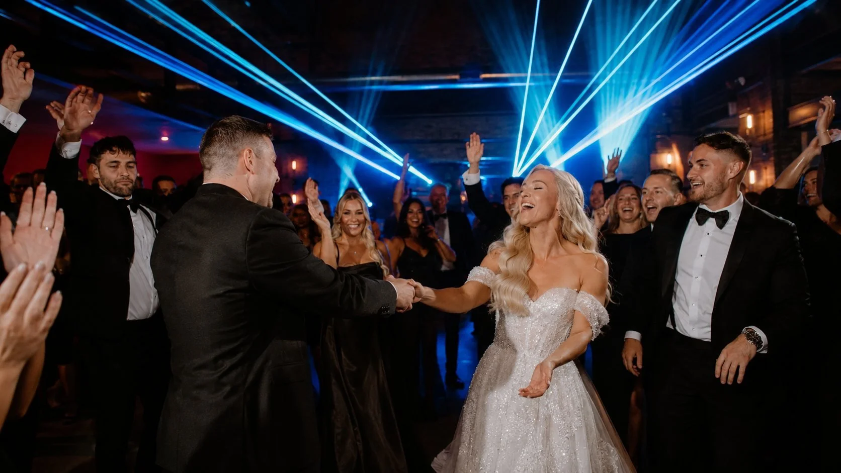 A bride and groom dancing at their wedding reception, surrounded by guests cheering and clapping under colorful laser lights.