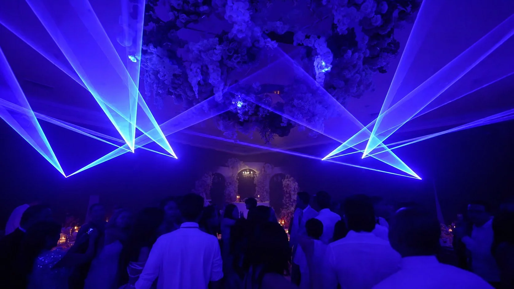 People dancing at a night event with blue laser lights and floral decorations.
