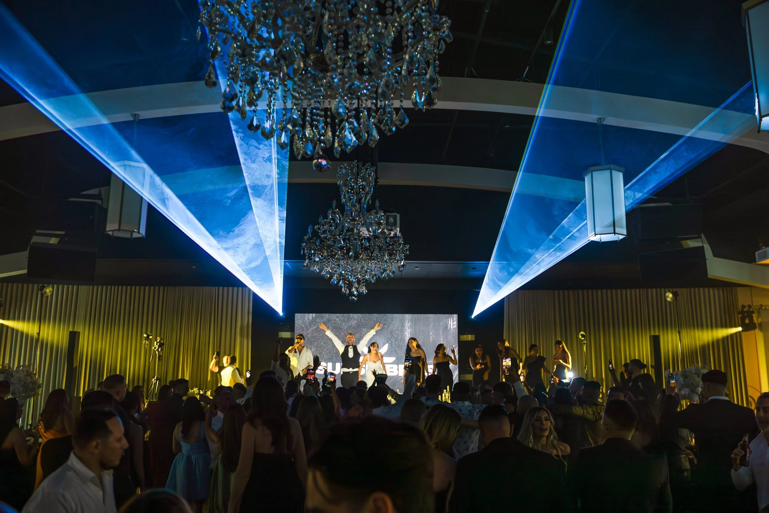 People dancing and singing on a stage at a party or celebration with chandeliers and blue laser lights overhead.