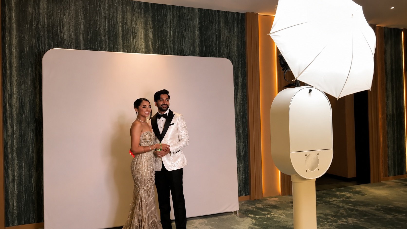 A man and woman dressed in formal attire standing close together for a photo in front of a white photo backdrop, with a large studio light and umbrella overhead.