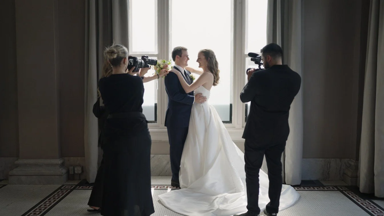 A bride and groom are embracing during their wedding ceremony while two videographers film the moment in front of a large window with curtains.