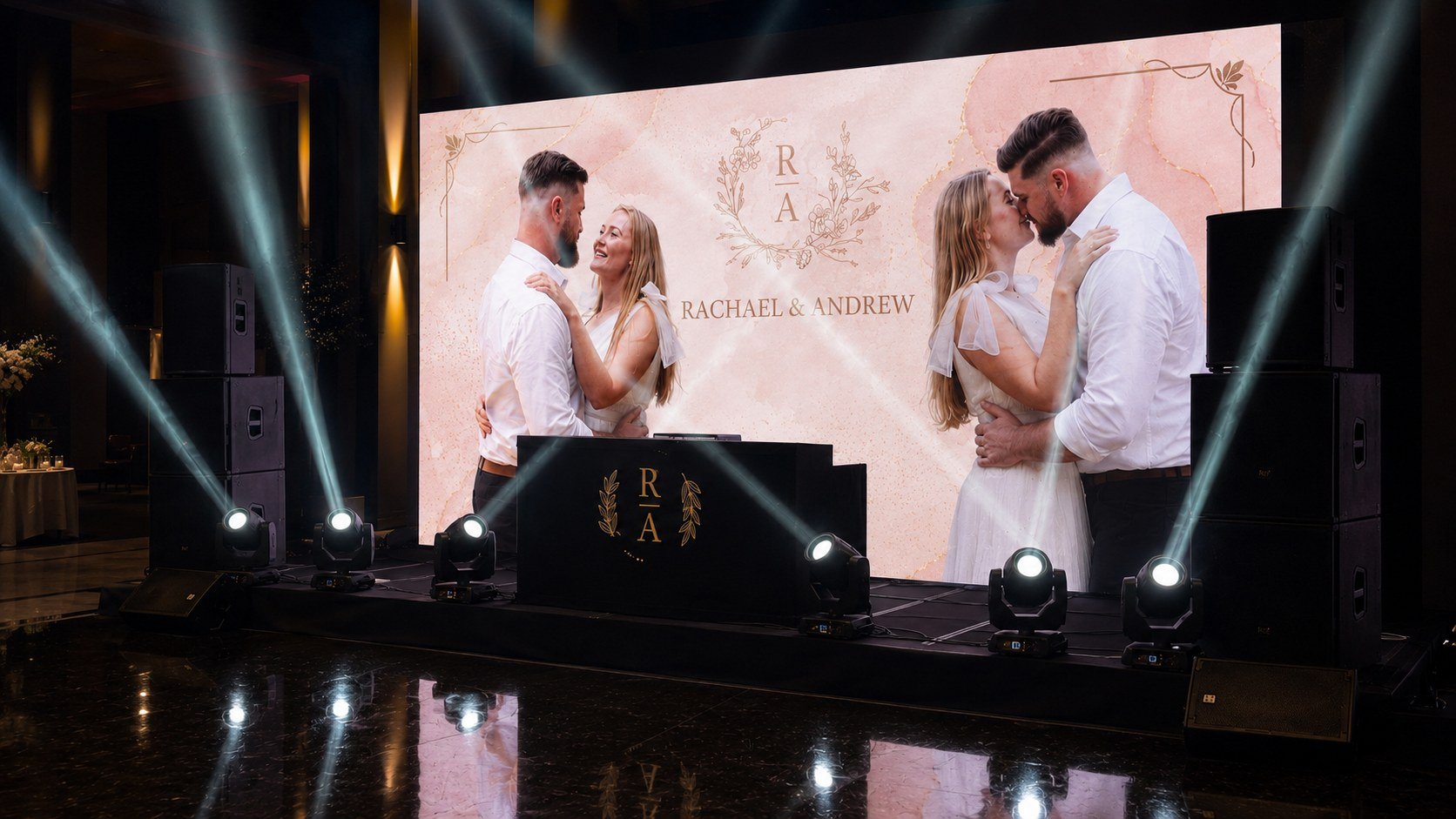 A newly married couple dances at their wedding reception with a large screen in the background displaying their names, Rachael and Andrew, and a monogram with the letters R and A surrounded by a floral design. The stage is decorated with lighting and speakers.