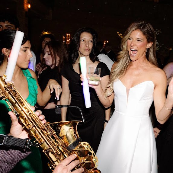 Woman in a white dress singing and dancing at a party, holding a drink, with a saxophone player nearby and other guests in the background.