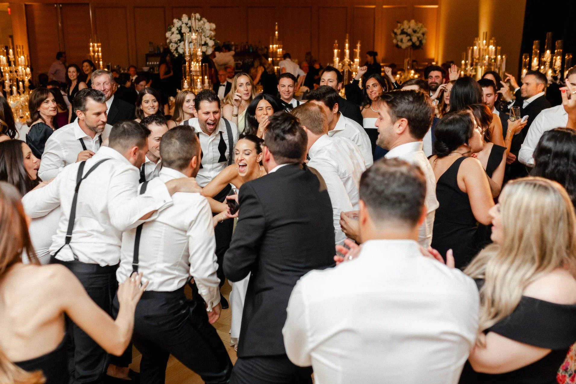 Guests dancing and celebrating at a formal event in a decorated ballroom with floral centerpieces and candlelit candelabras.