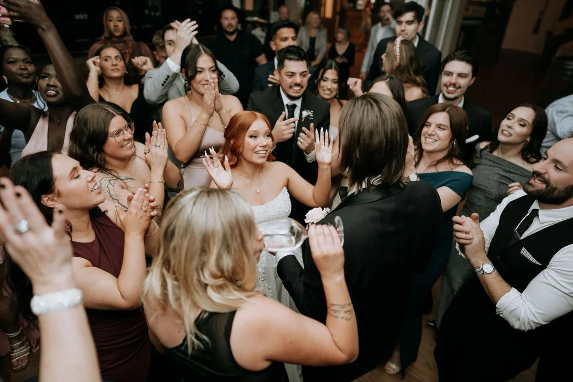 A group of people gathered closely together at a celebration, with some smiling and clapping, at a wedding reception. The bride with red hair in a white dress is in the center, smiling with her hands raised, and surrounded by friends and family.