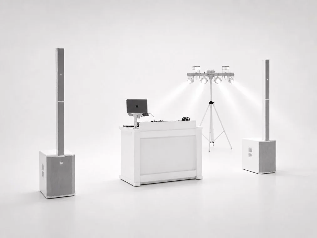 A DJ setup in a minimalistic white studio with two tall speakers, a DJ booth with a computer, and a light rig with four spotlights.