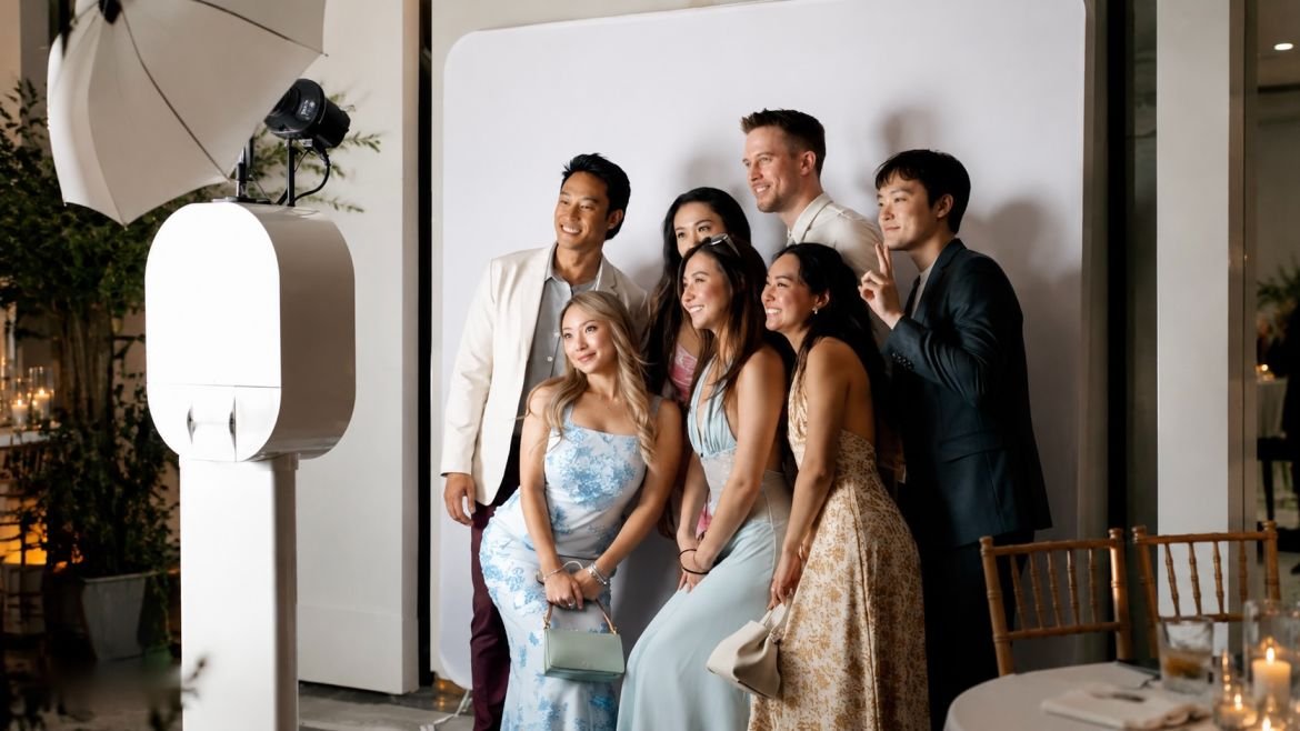 Group of seven people posing for a photo at a party or celebration, standing in front of a white backdrop, with professional lighting setup nearby.