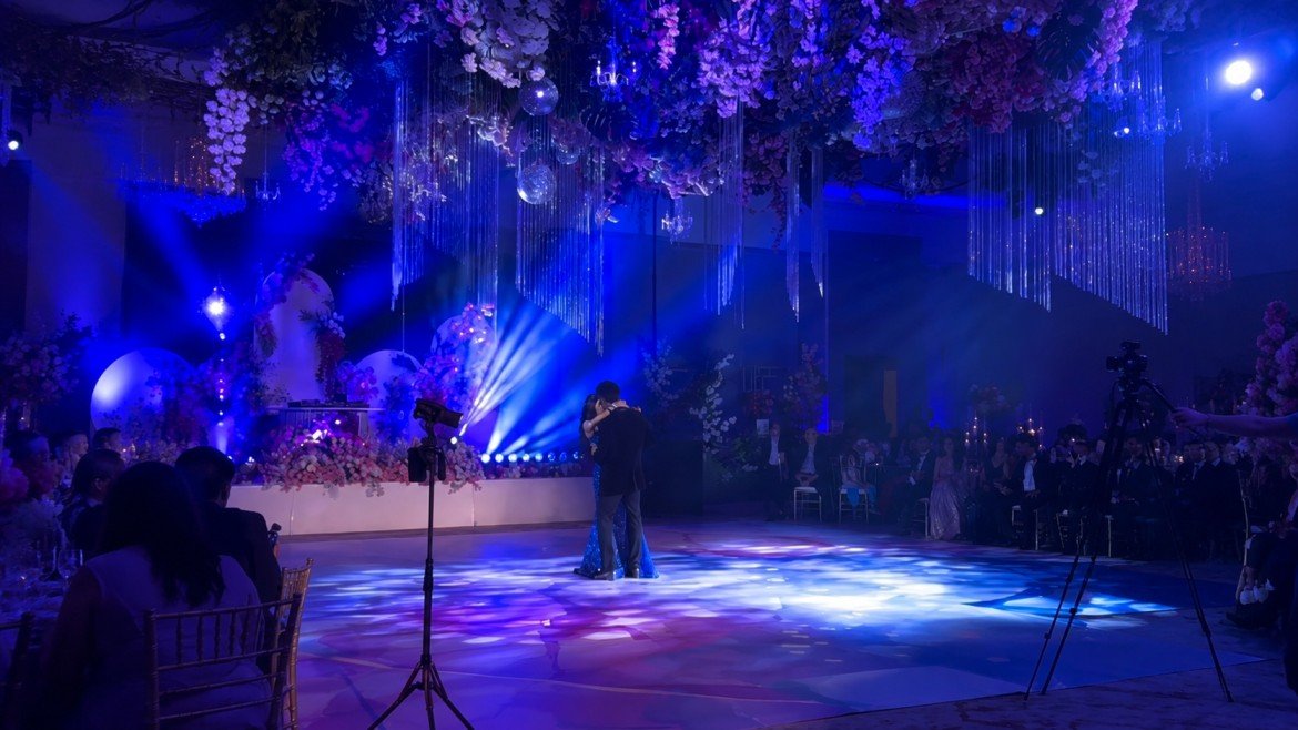 A couple is dancing center stage at a decorated wedding reception with purple and blue lighting, hanging flowers, and chandeliers, surrounded by seated guests.