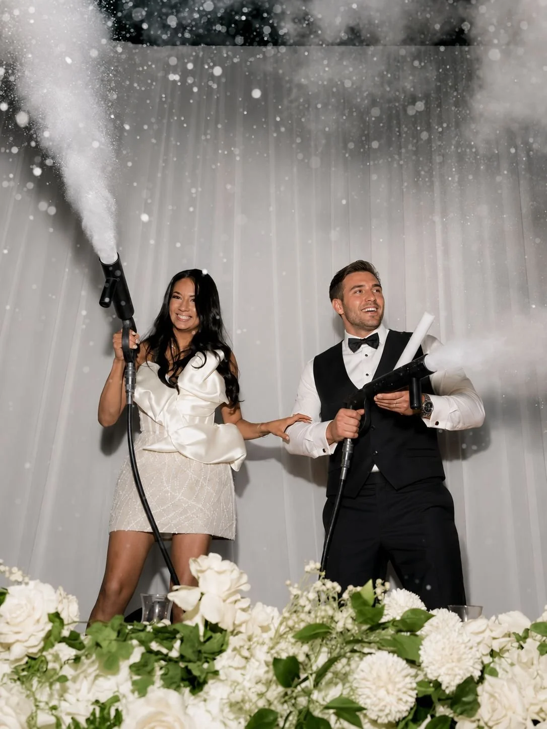 A couple dressed in formal attire is celebrating at a wedding, spraying champagne and smiling, surrounded by white flowers.