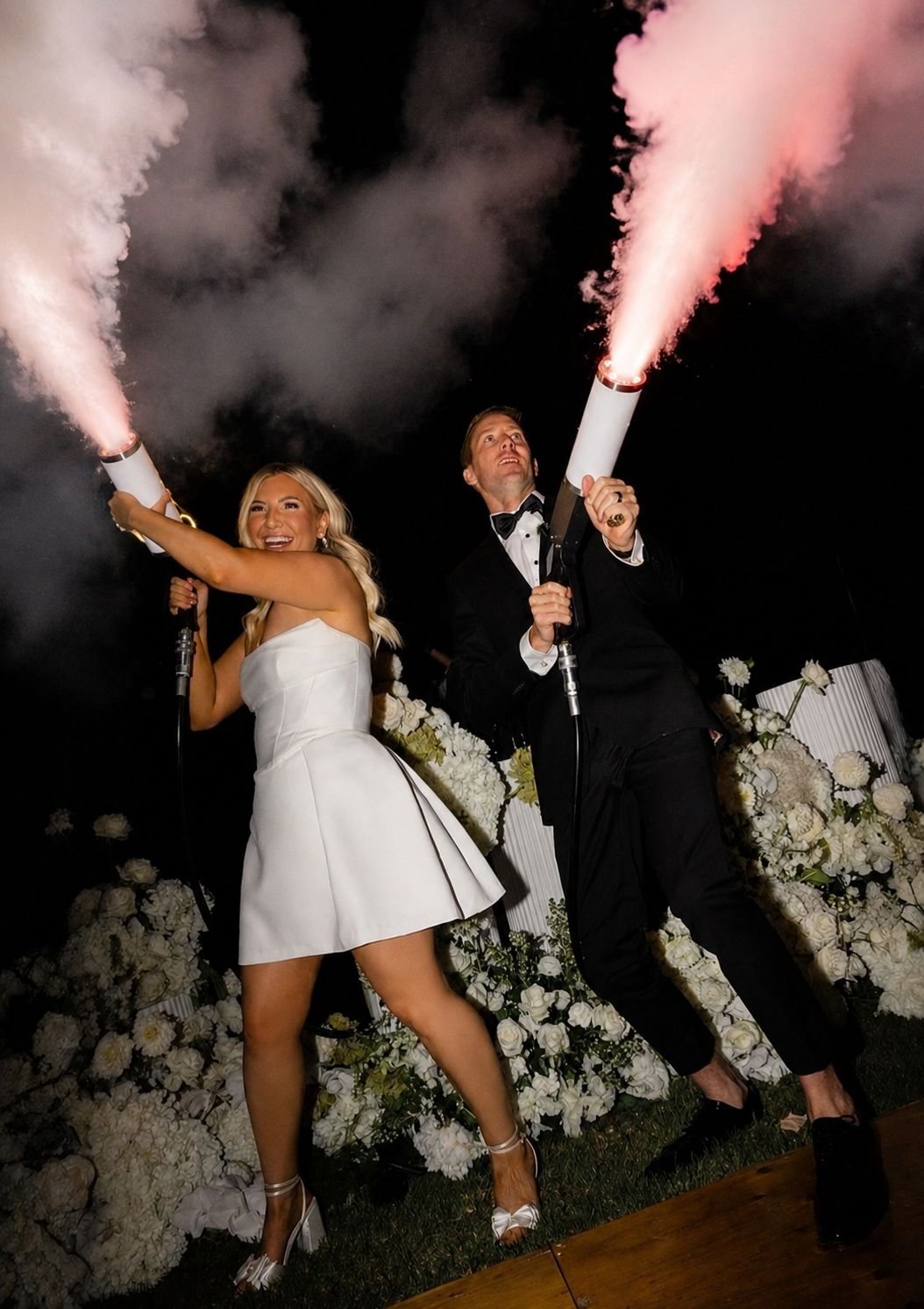 Two people, a woman in a white dress and a man in a tuxedo, holding and firing party poppers that release pink smoke at nighttime, with a background of white floral arrangements.