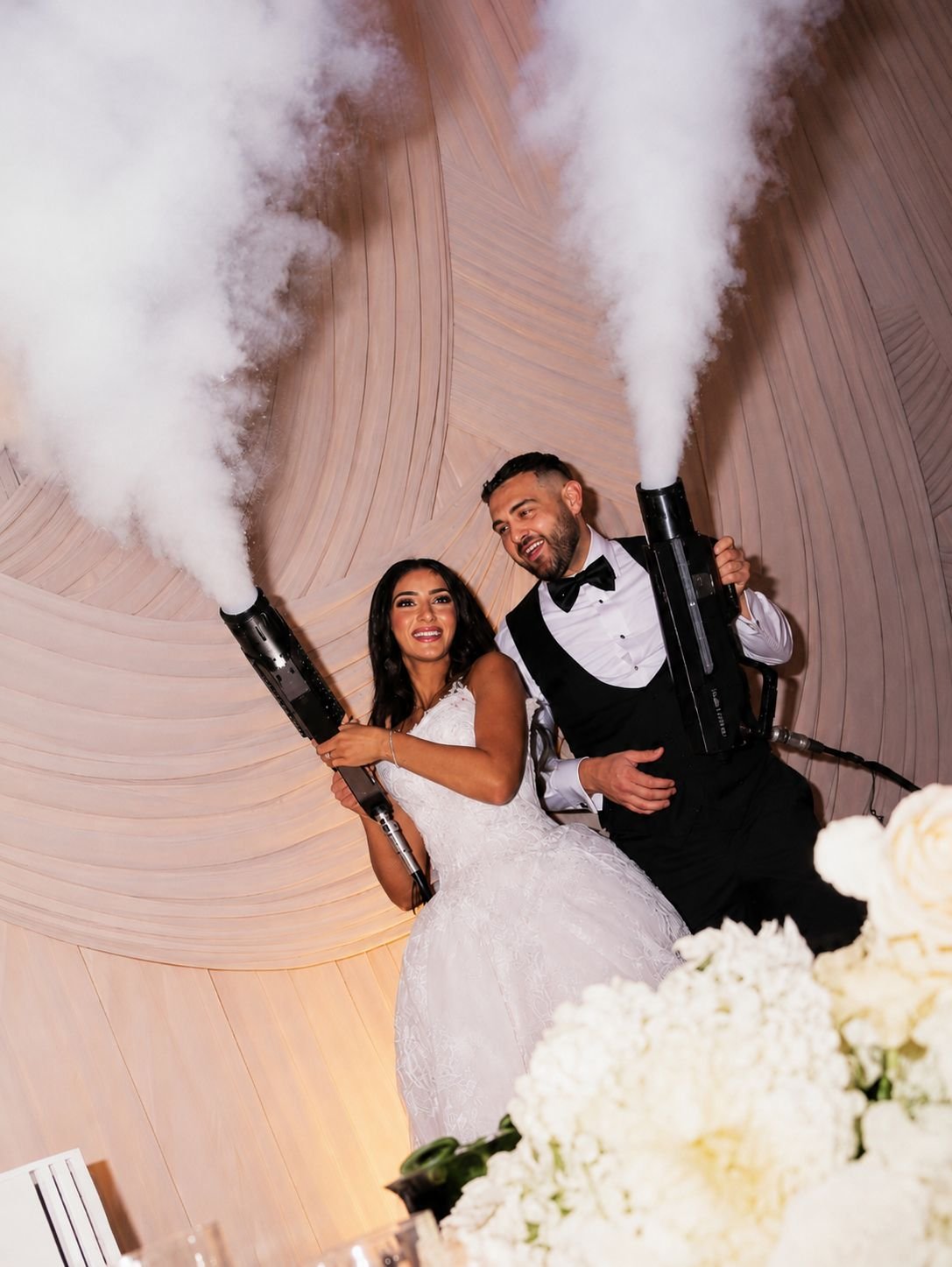 A bride and groom holding smoke cannons at their wedding reception, with the bride in a wedding dress and the groom in a tuxedo, smiling.