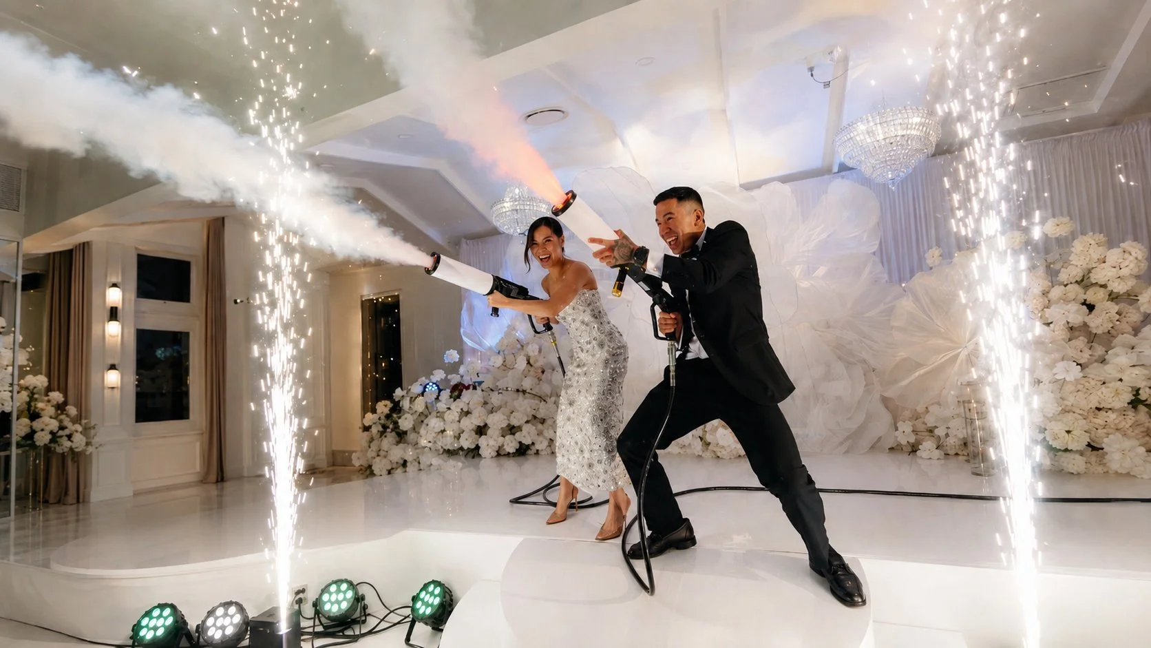 A couple celebrating at their wedding reception, wearing formal attire, holding party cannons that shoot confetti and sparks, surrounded by white floral decorations and sparklers, with joyful expressions.