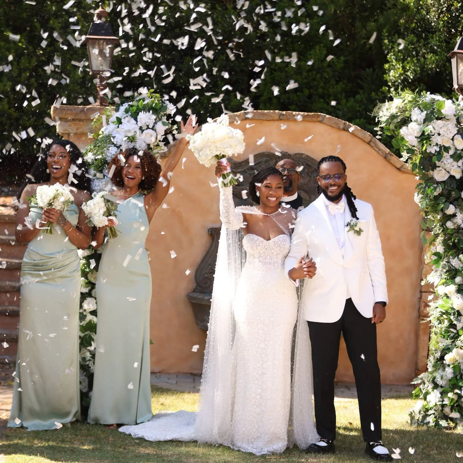 A wedding celebration with a bride, groom, and three bridesmaids. The bride and groom are holding hands, smiling, and standing under a floral arch with white flowers and greenery. The bridesmaids are holding bouquets and throwing white confetti. The setting appears to be outdoors with greenery and a stone wall in the background.