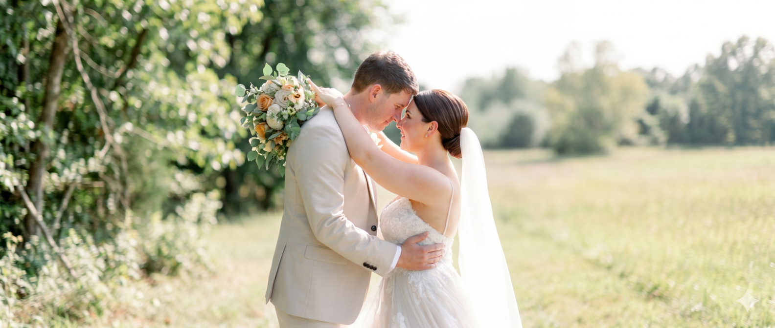 A bride and groom embrace outdoors in a field on their wedding day, with the bride holding a bouquet of flowers and they are touching foreheads.