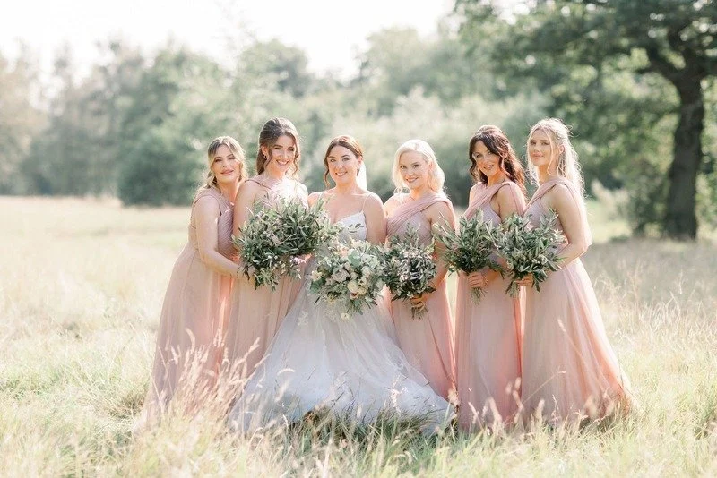 A bride in a white wedding gown standing with five bridesmaids in pink dresses outdoors in a field with trees in the background, holding bouquets of flowers.