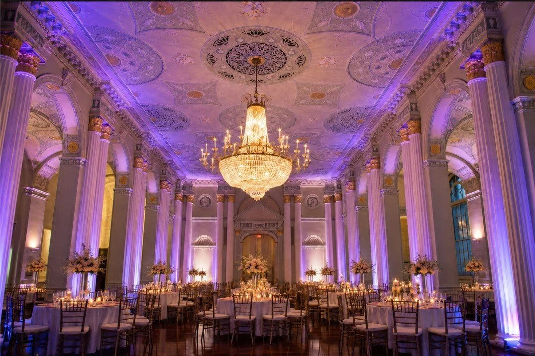 Elegant ballroom decorated for a formal event with tall columns, a large chandelier, purple and warm lighting, round tables with white tablecloths, candles, and floral centerpieces.