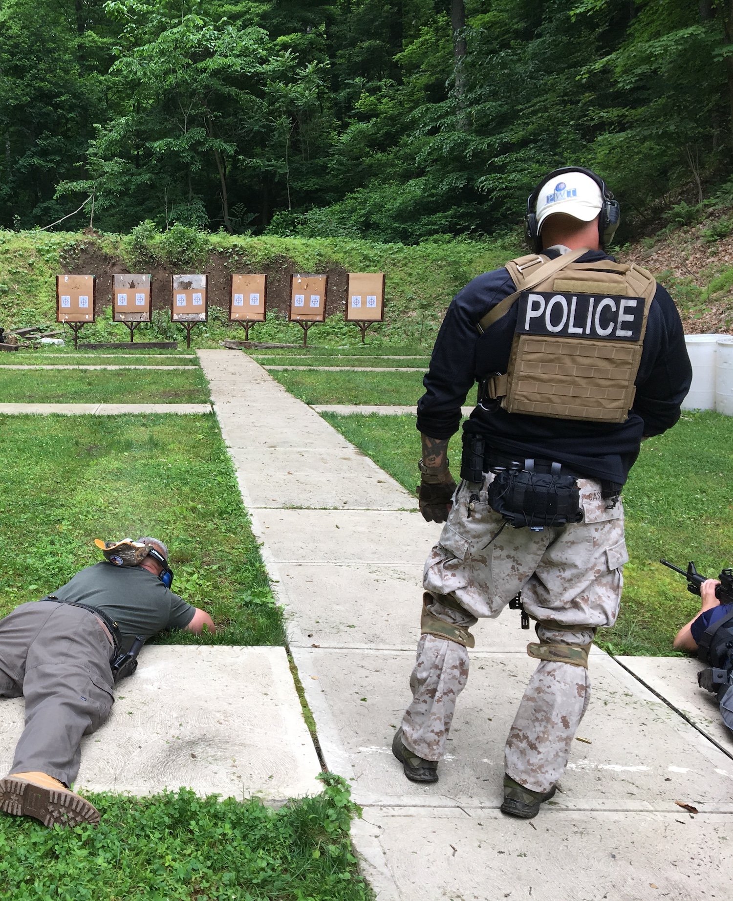 A police officer in camouflage pants and a tan vest with 'POLICE' on the back observes a shooting range with several targets set up on a grassy hillside. Two individuals are lying on the ground, aiming their firearms at the targets, while another person is crouched nearby. The area is surrounded by green trees and grass.