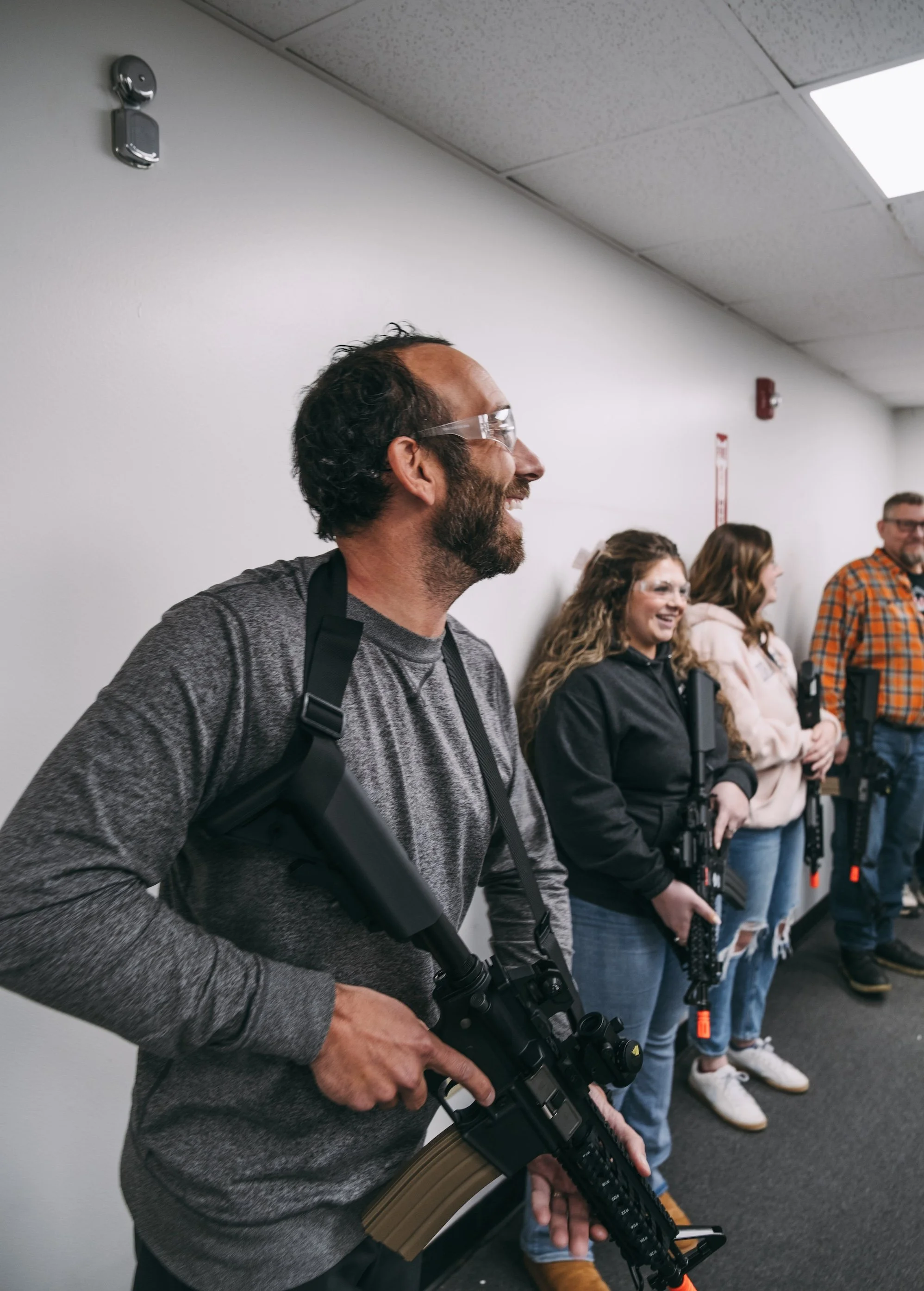 A group of people standing in a line against a white wall, each holding a rifle, with the focus on a man in the foreground wearing safety glasses and a gray shirt, smiling.