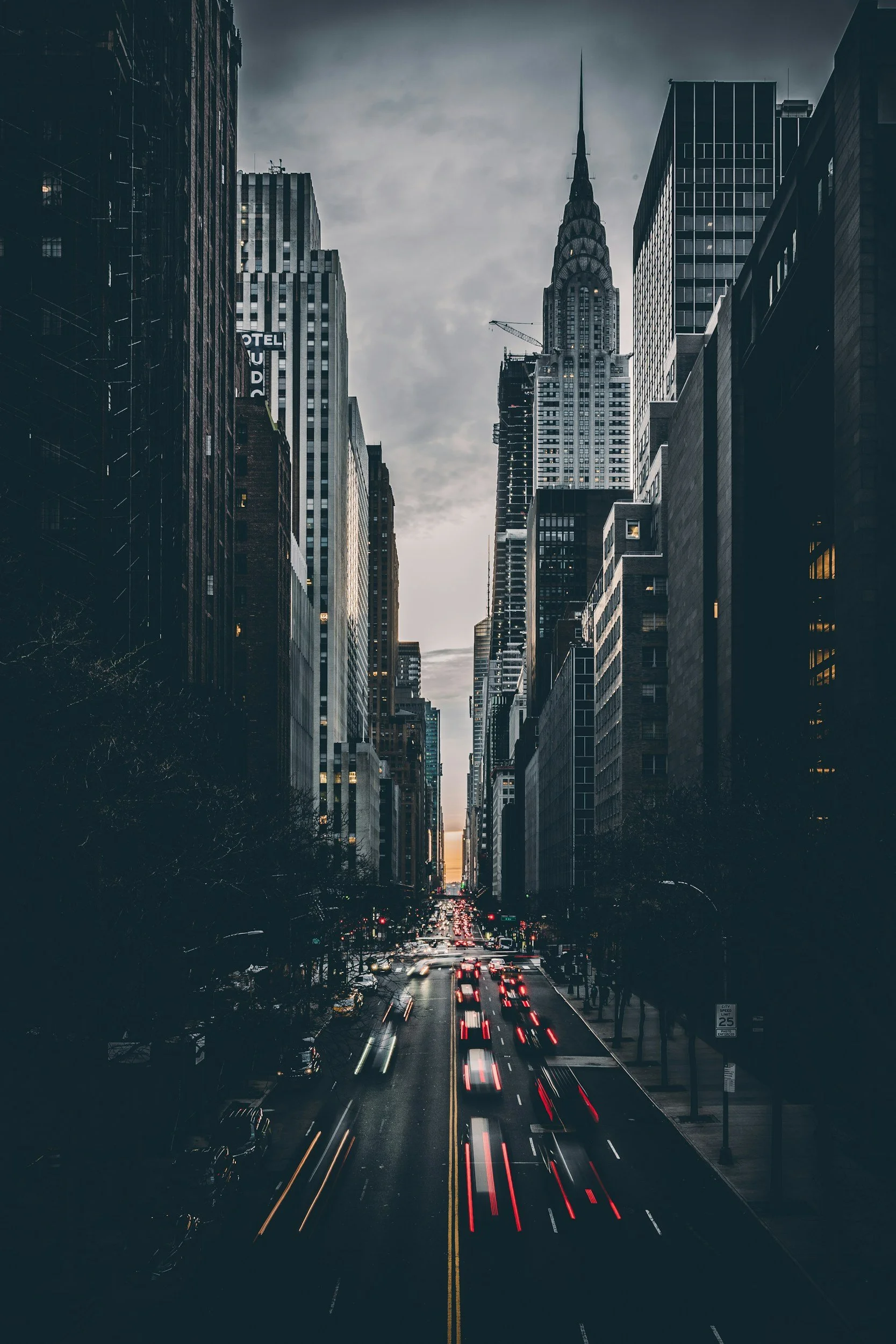 City street in New York City at dusk, with tall skyscrapers on both sides, a cloudy sky, and vehicle headlights creating light trails on the road.