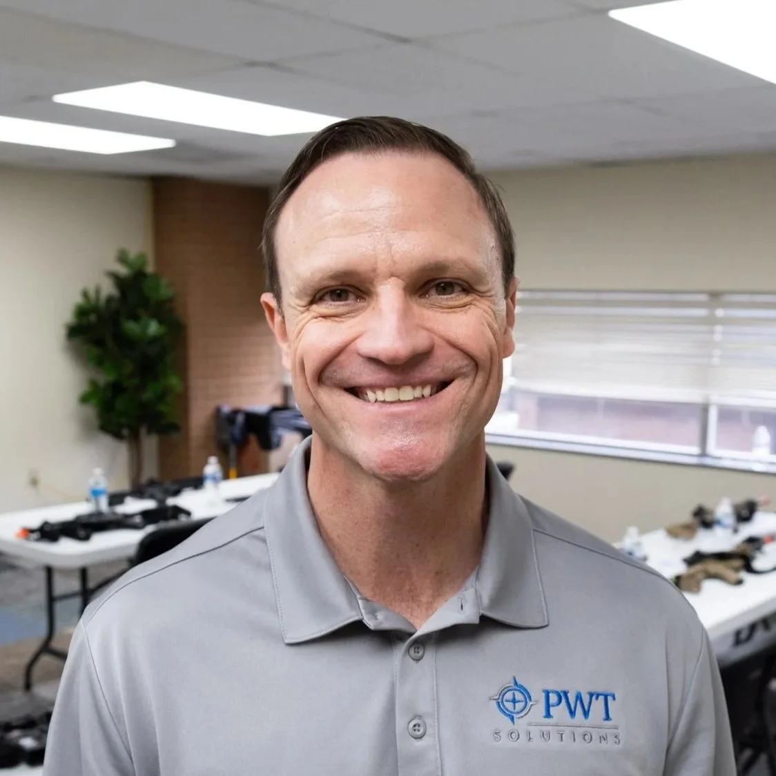 A smiling man in a gray polo shirt with PWT Solutions logo, standing in a conference room with tables, water bottles, and some equipment in the background.