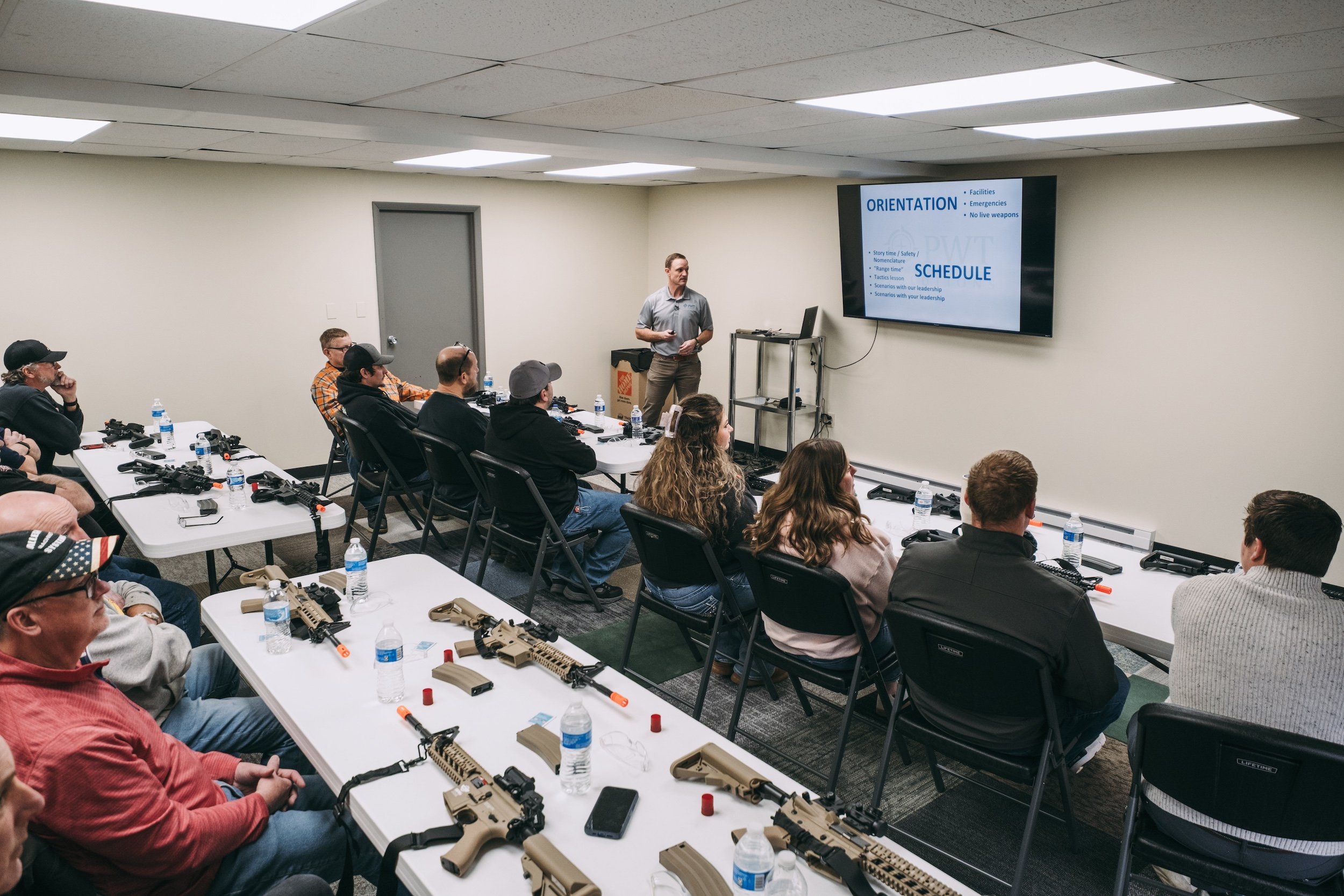 A classroom training session with students seated at tables equipped with firearms and magazines. An instructor stands at the front near a large screen displaying course information. Most attendees are listening attentively, with some taking notes.