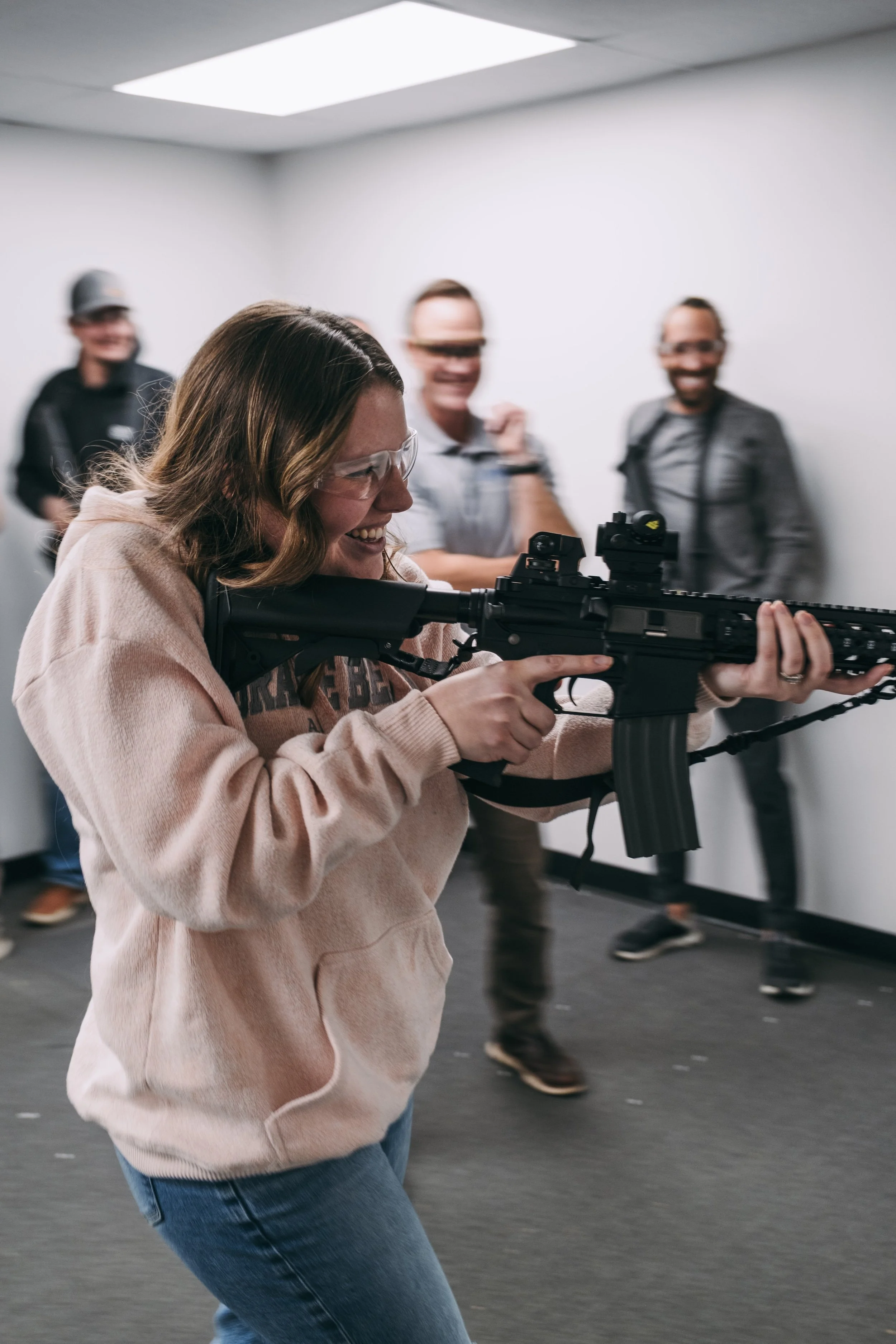 A woman in a beige hoodie aiming and holding a rifle with a scope in an indoor shooting range, with three smiling men in the background.