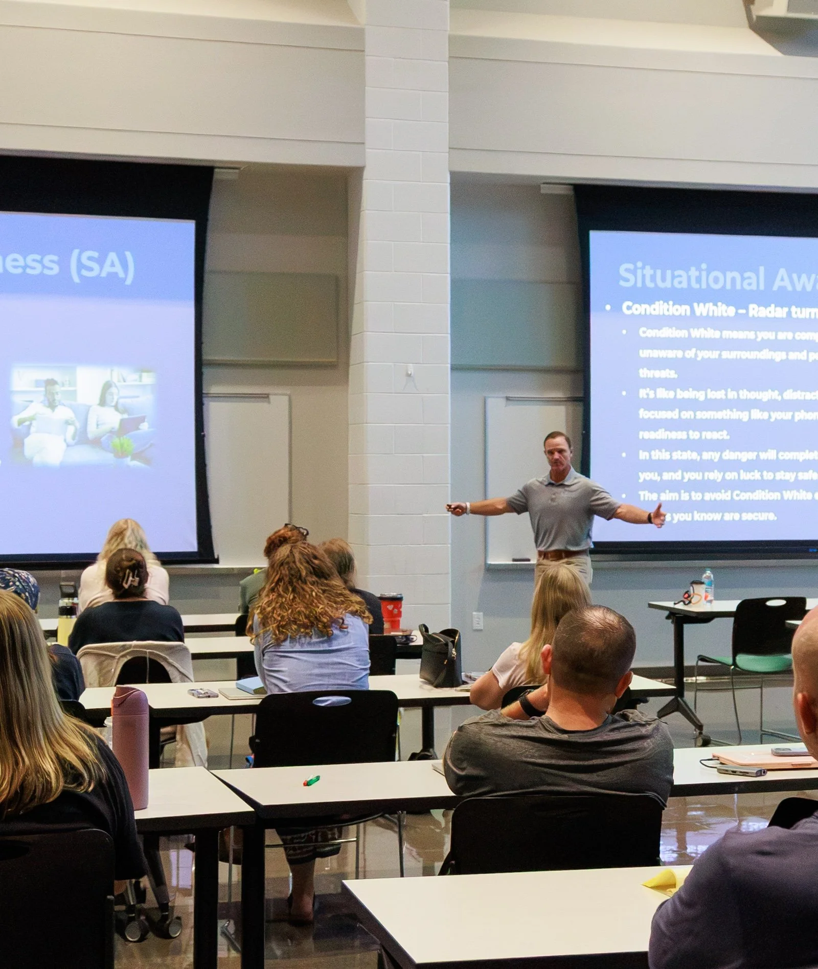 A man giving a presentation at a conference or classroom, standing in front of two large screens with presentation slides, with students or attendees seated at desks facing him.
