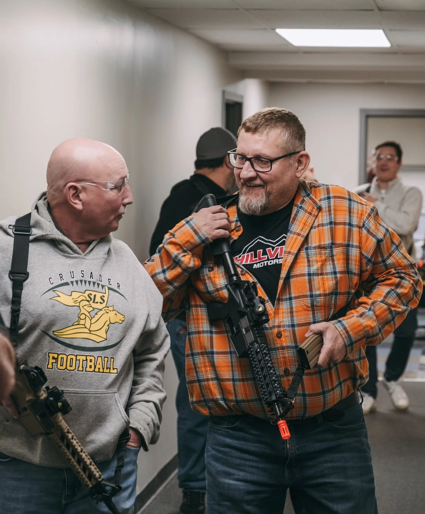 Two men indoors, one with a gray hoodie and the other in an orange plaid shirt, are holding rifles and smiling, with several other people in the background.