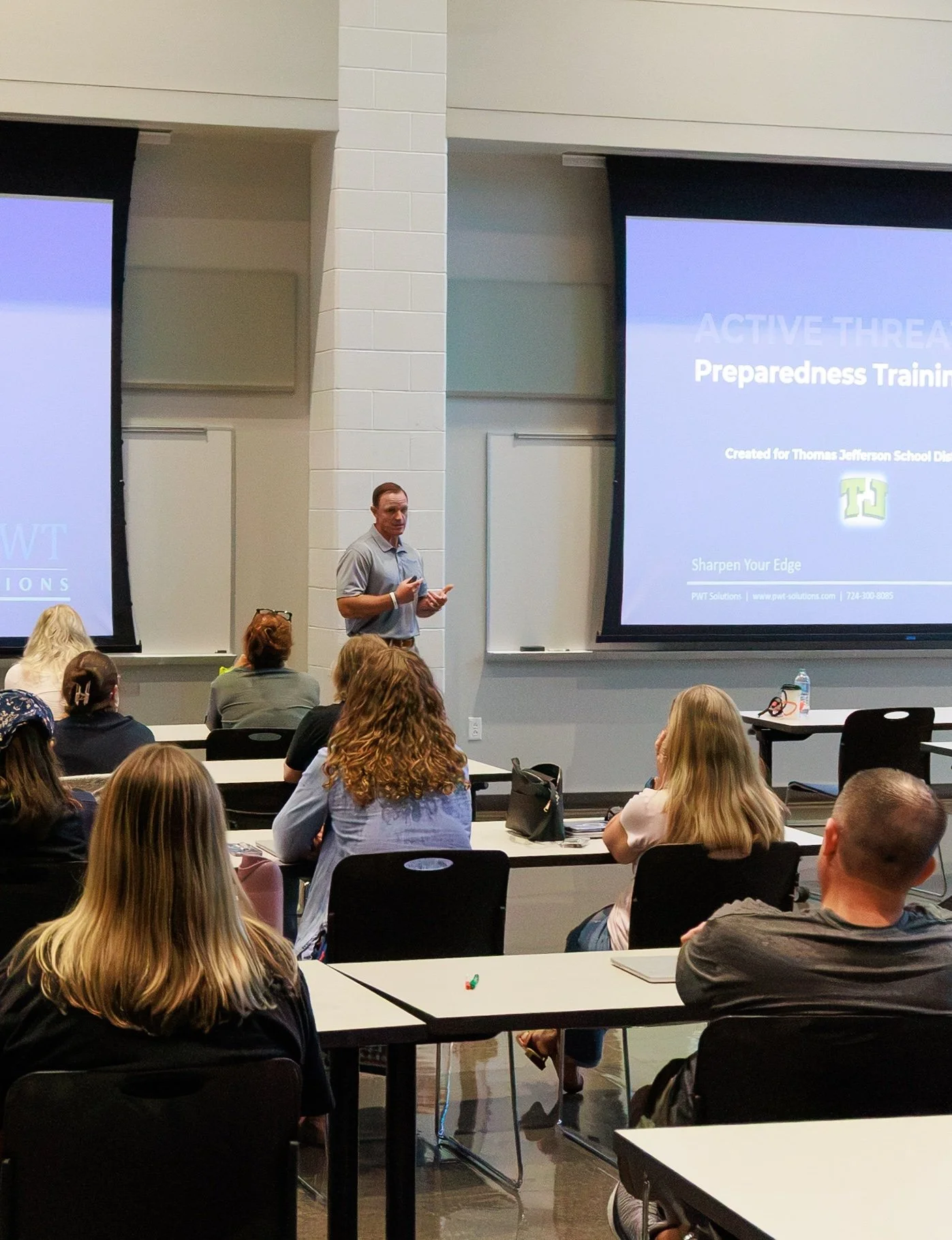 A man giving a presentation in a classroom or conference room with an audience of adults. The slide on the screen behind him reads 'ACTIVE THREAT' and 'Preparedness Training' and includes a logo and contact information for PWET Solutions.