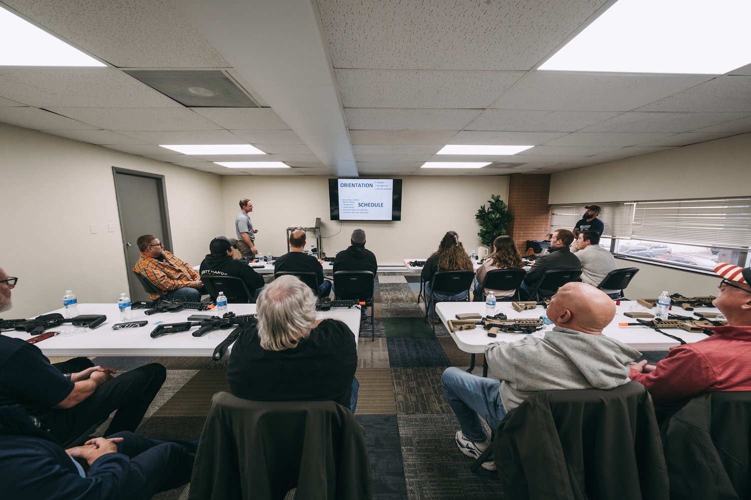A classroom or training room with a group of people sitting around tables, some with firearms on the table. A man is standing at the front near a large screen displaying training schedule details. There are windows with blinds on the right side and a brick accent wall with a plant. The people appear to be participating in a safety or training seminar.