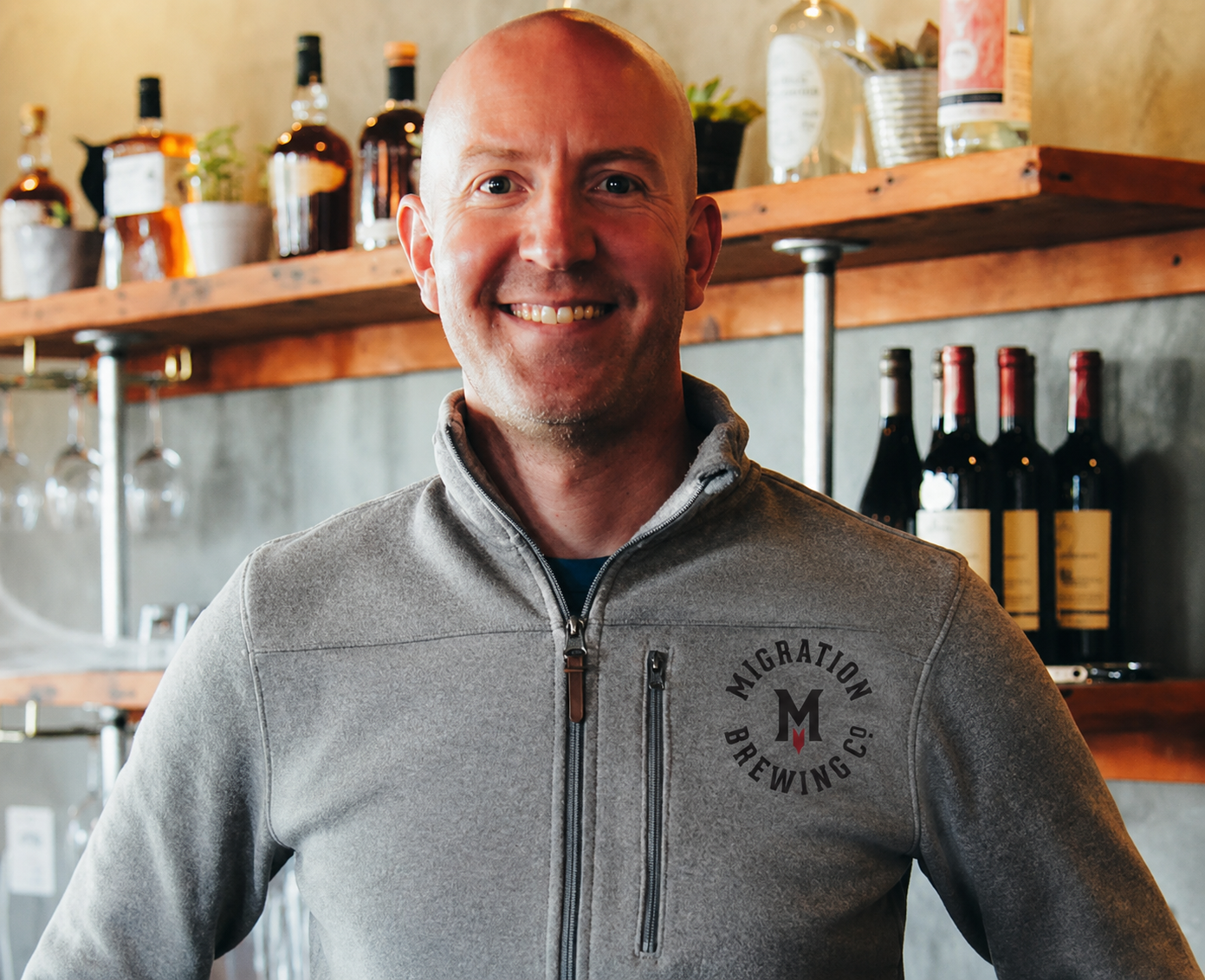 Migration Brewing Company CEO, wearing a gray zip-up jacket with 'Migration Brewing Co' logo stands inside a brewery, with shelves of bottles and glasses behind him.