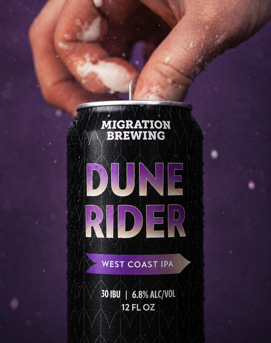 Product hero shot.  Social content.  A black can of Dune Rider West Coast IPA from Migration Brewing with water droplets, held by a hand, against a dark purple background with a person’s fingers and some foam or powder on them.