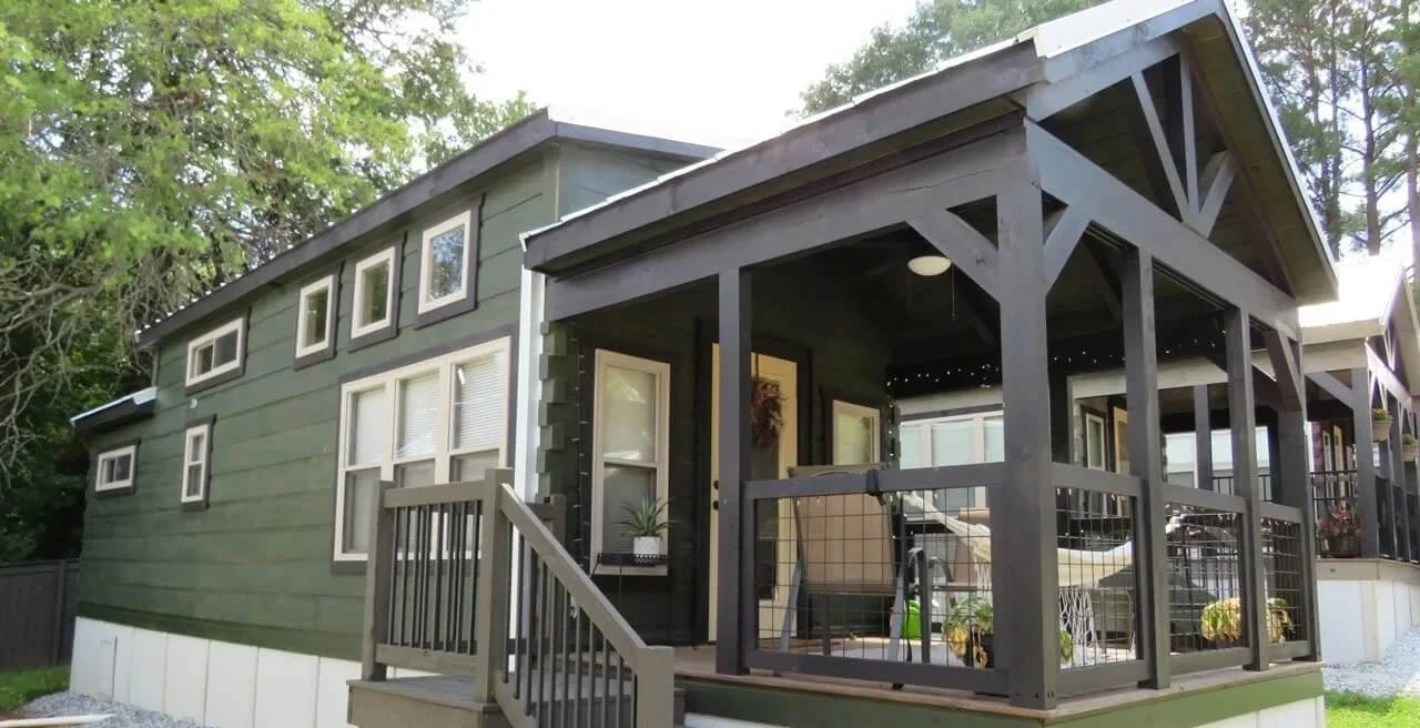 A modern green house with a covered porch, multiple windows, and a surrounding outdoor space with chairs and decorations.