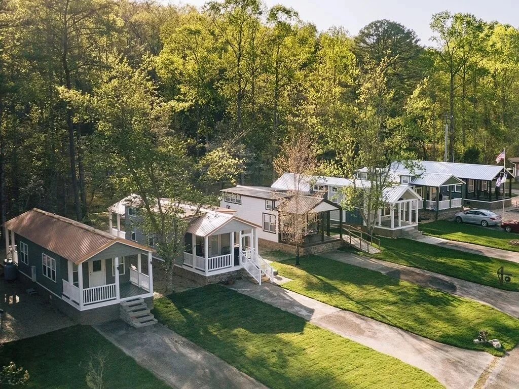 A row of small American suburban houses with green lawns, driveways, and trees, with a wooded area in the background.