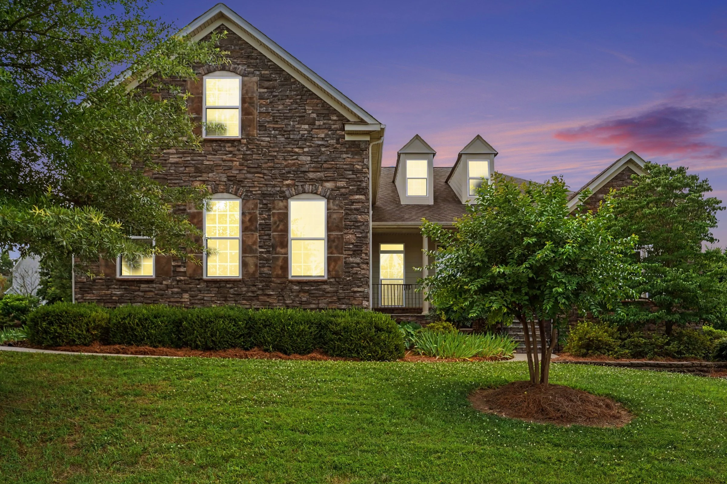 A two-story house with a brick exterior, multiple windows with yellow light inside, and a gabled roof, surrounded by green shrubs and trees during dusk.