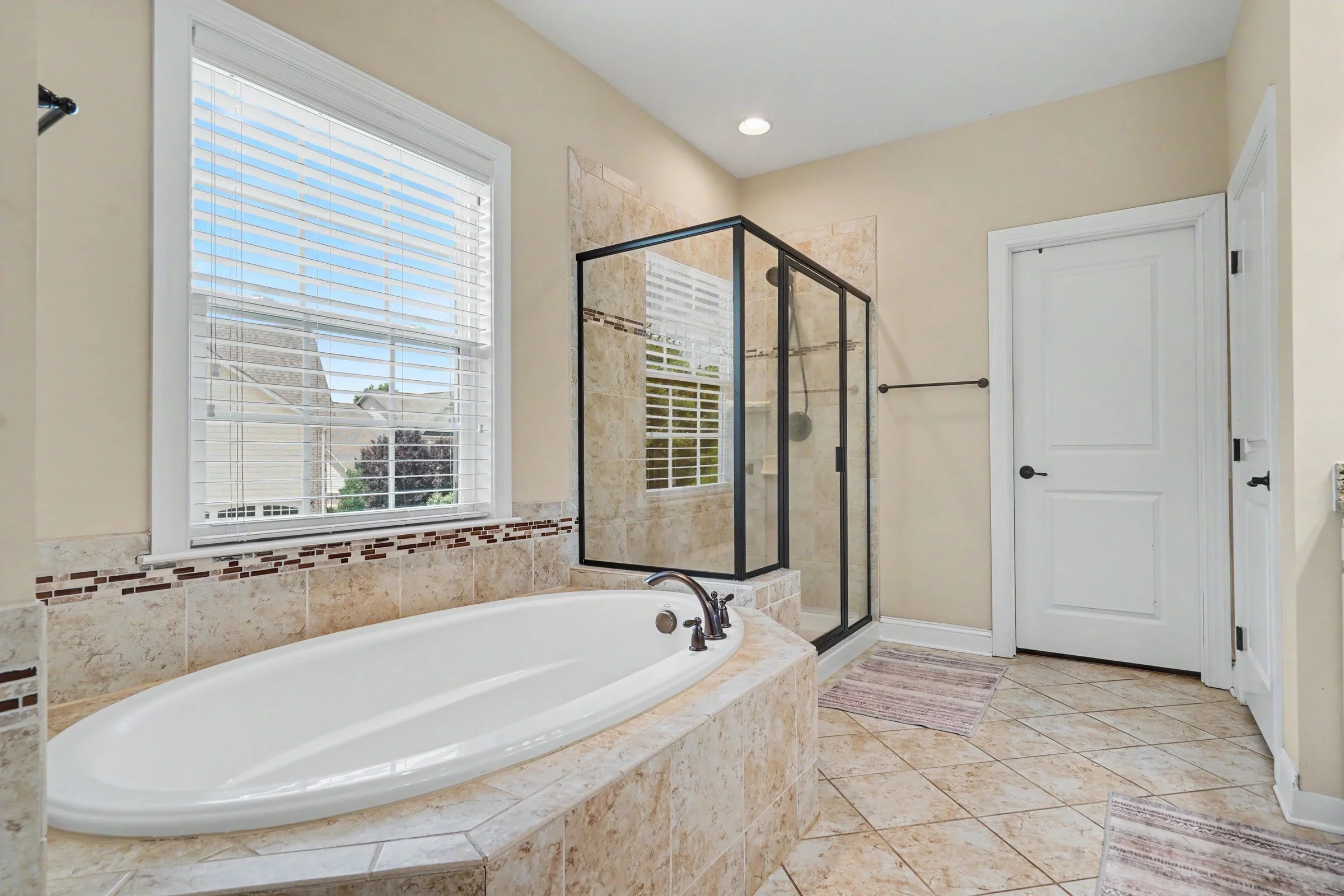 Bathroom with a large bathtub, a walk-in shower with glass walls, a window with blinds, and a door, all featuring beige tiles and trim.