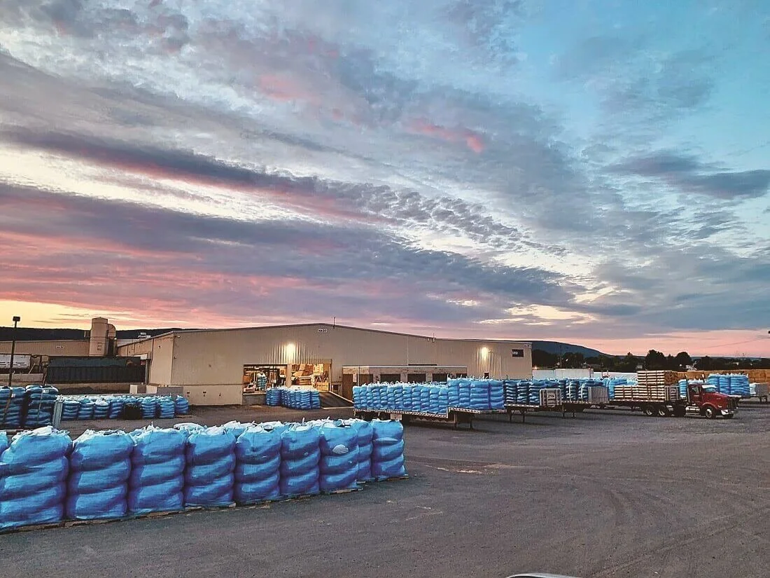 The image shows an outdoor warehouse area during sunset with large stacks of blue bags and pallets, a warehouse building with open door, a red truck, and a colorful sky with clouds.