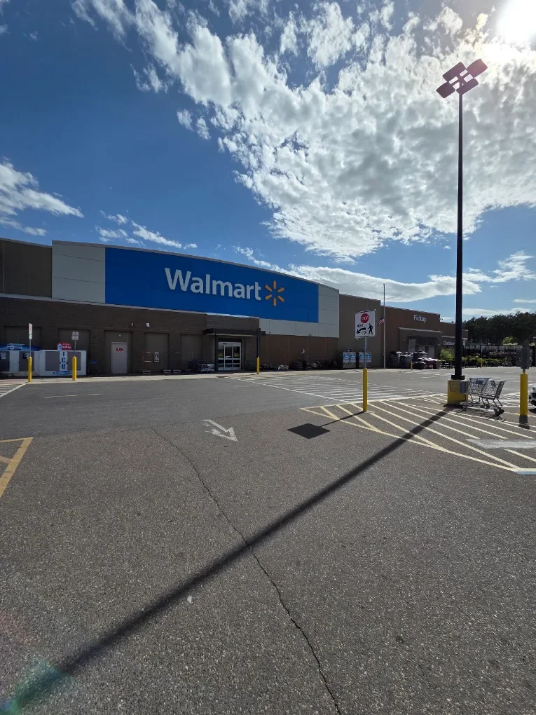 Exterior view of a Walmart store with a parking lot in the foreground, a shopping cart, and a light pole. The sky is partly cloudy with sunlight.