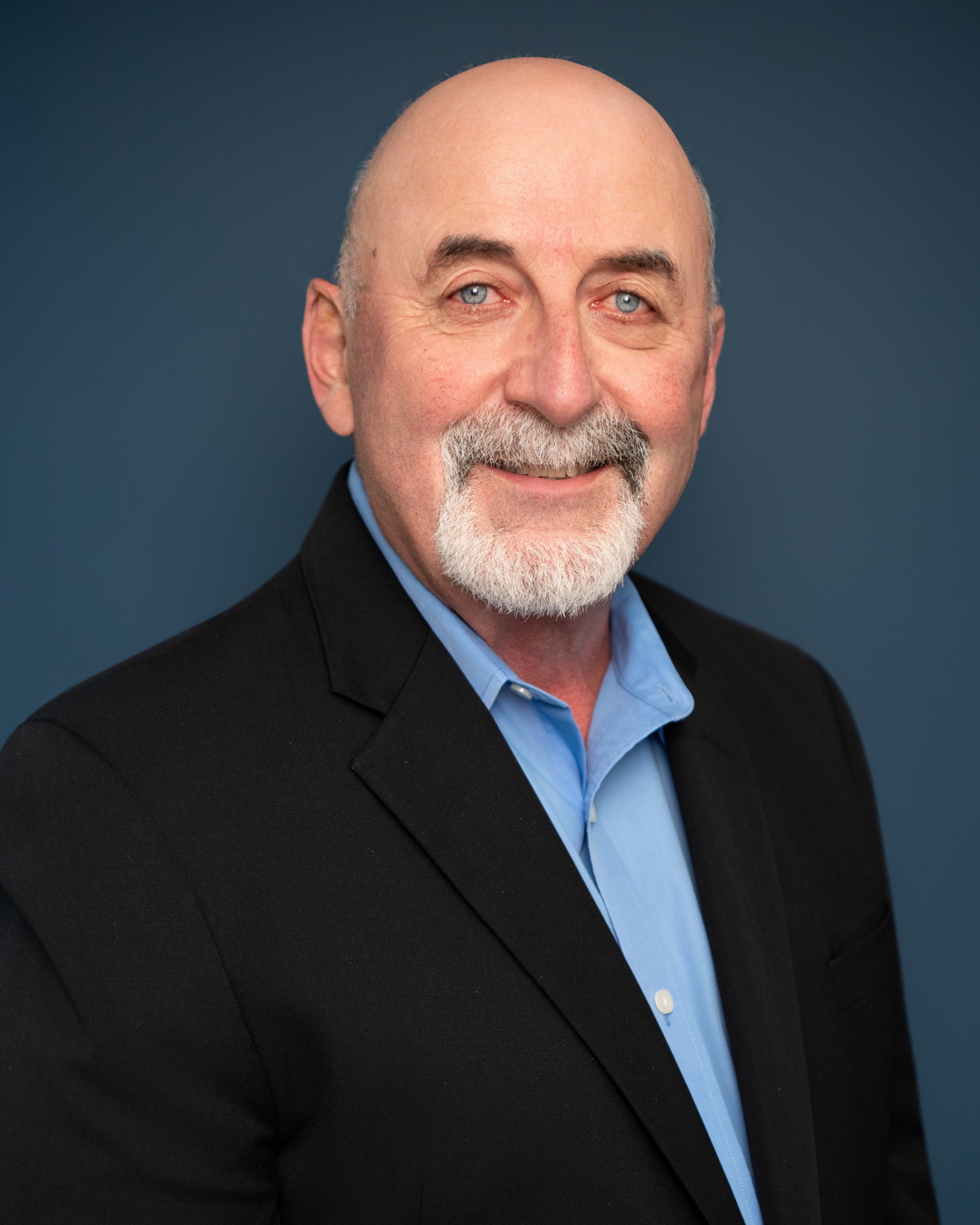 Close-up portrait of a smiling man with a bald head, blue eyes, and a gray beard. He is wearing a dark blazer and a light blue dress shirt, set against a blue background.