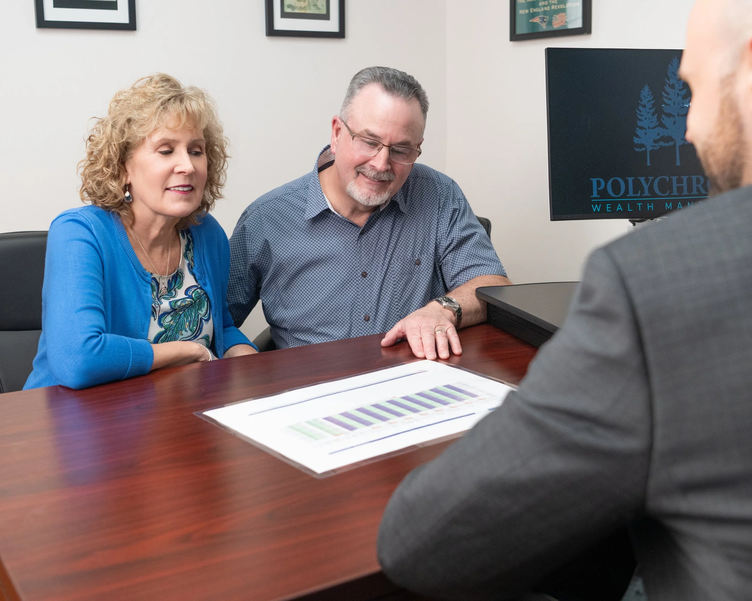Three people sitting around a conference table, looking at documents and discussing. A woman on the left wears a blue cardigan, a man in the middle has glasses and a checkered shirt, and another person on the right is partially visible.