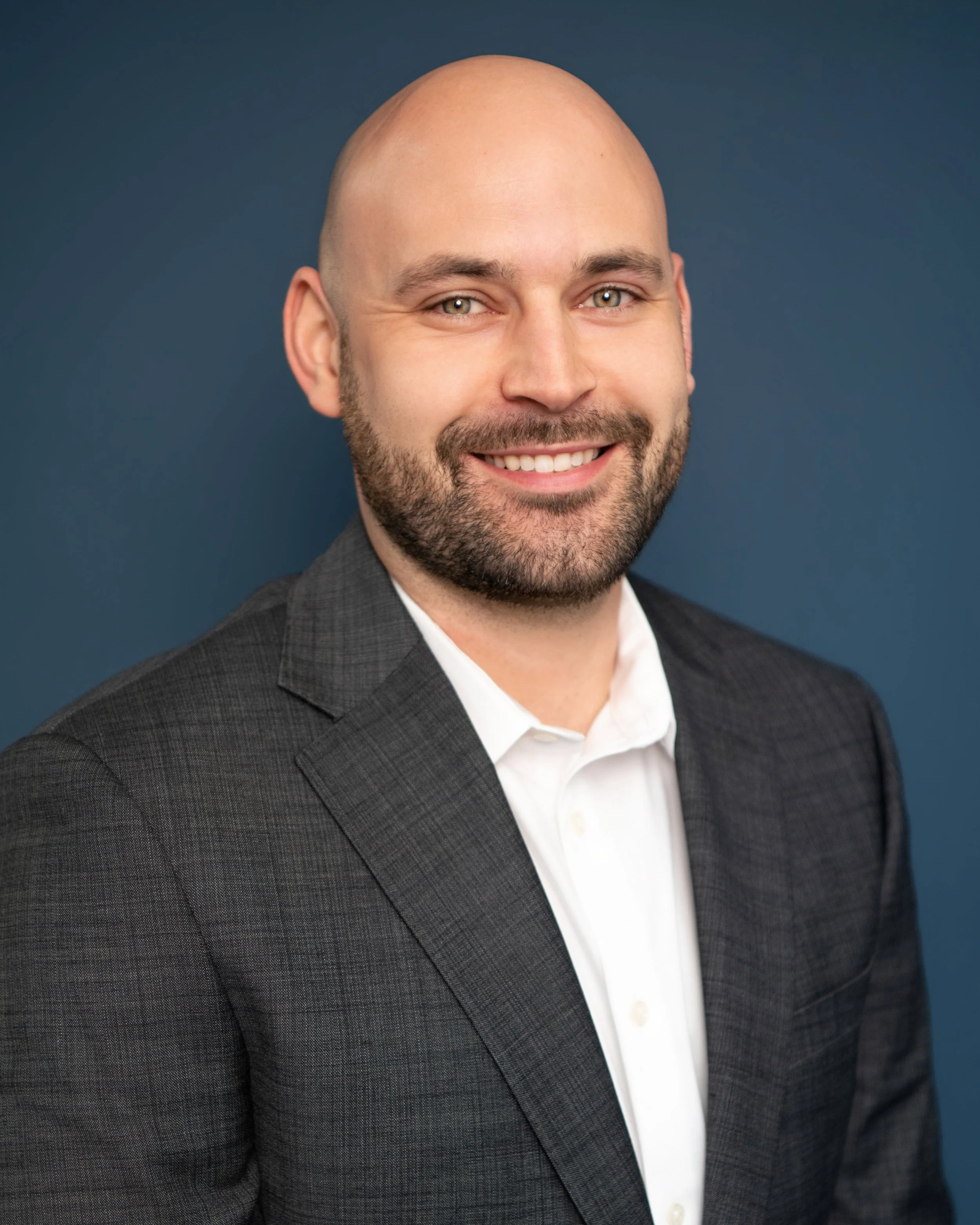Close-up portrait of a smiling bald man with a beard, wearing a dark gray checkered suit jacket and a white shirt, against a solid dark blue background.