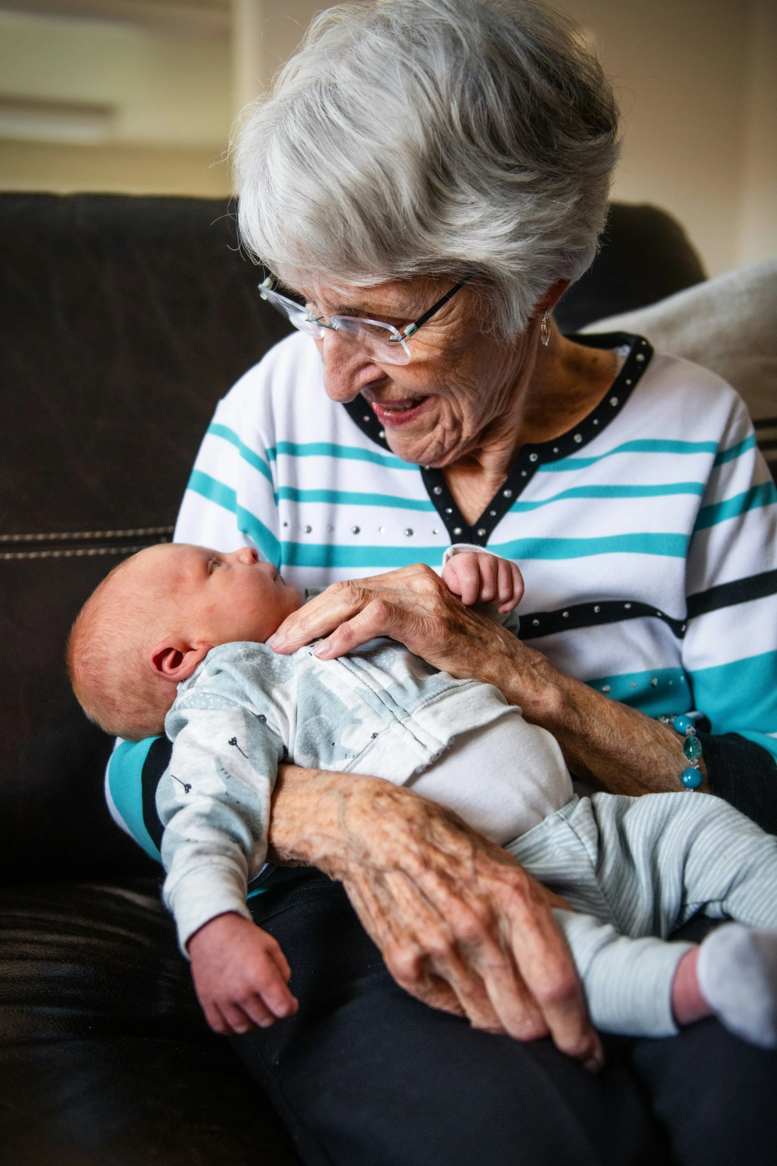 An elderly woman with gray hair and glasses is sitting on a black couch, holding a baby in her arms and looking down at the baby with affection.
