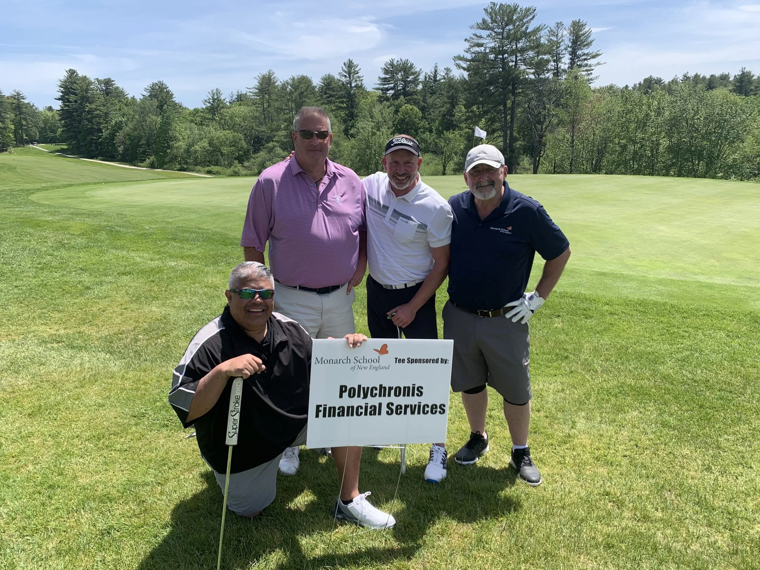 Four men posing on a golf course, with three standing and one kneeling in the front, holding a sign with business names, in a lush green landscape with trees and a blue sky.